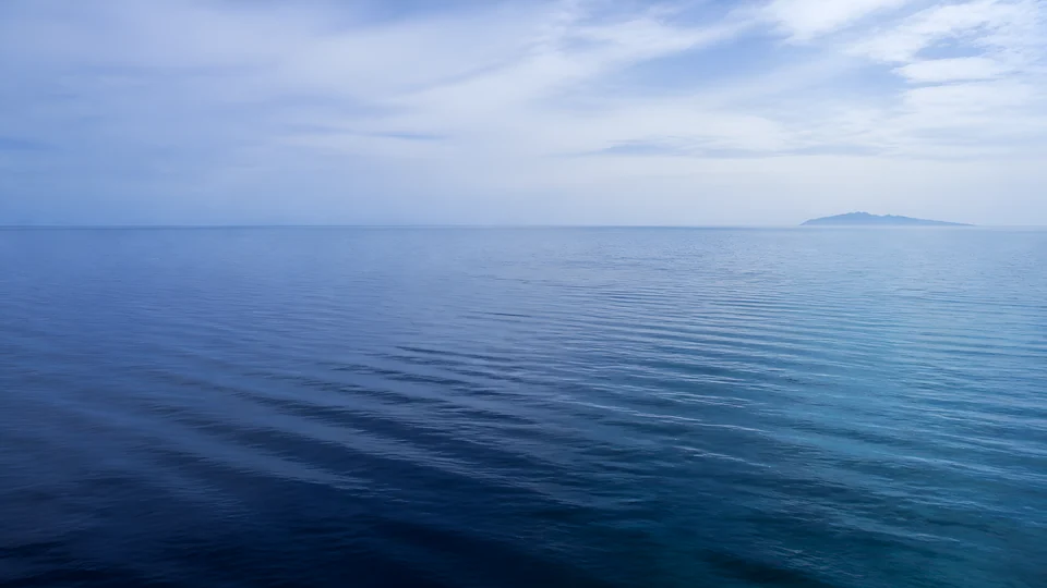A bright-overcast Pacific lagoon shifts from cobalt calm to muted blue-green beneath a distant island horizon.