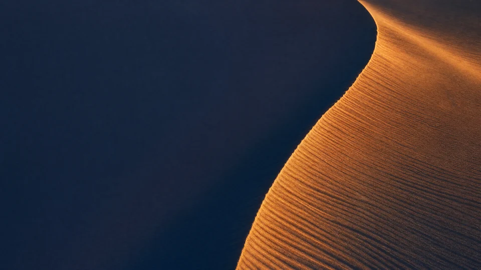 A crescent dune holds copper daylight and indigo moon-shadow on opposite faces of one razor ridge.